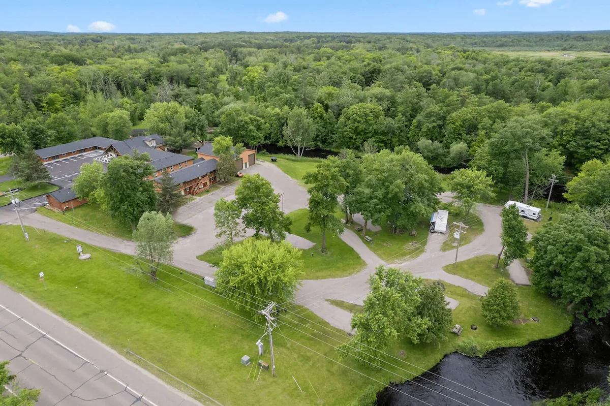 Aerial view of a wooded area with buildings, winding roads, parked RVs, a small river, and a main road running along the edge of the property, all surrounded by lush green trees.