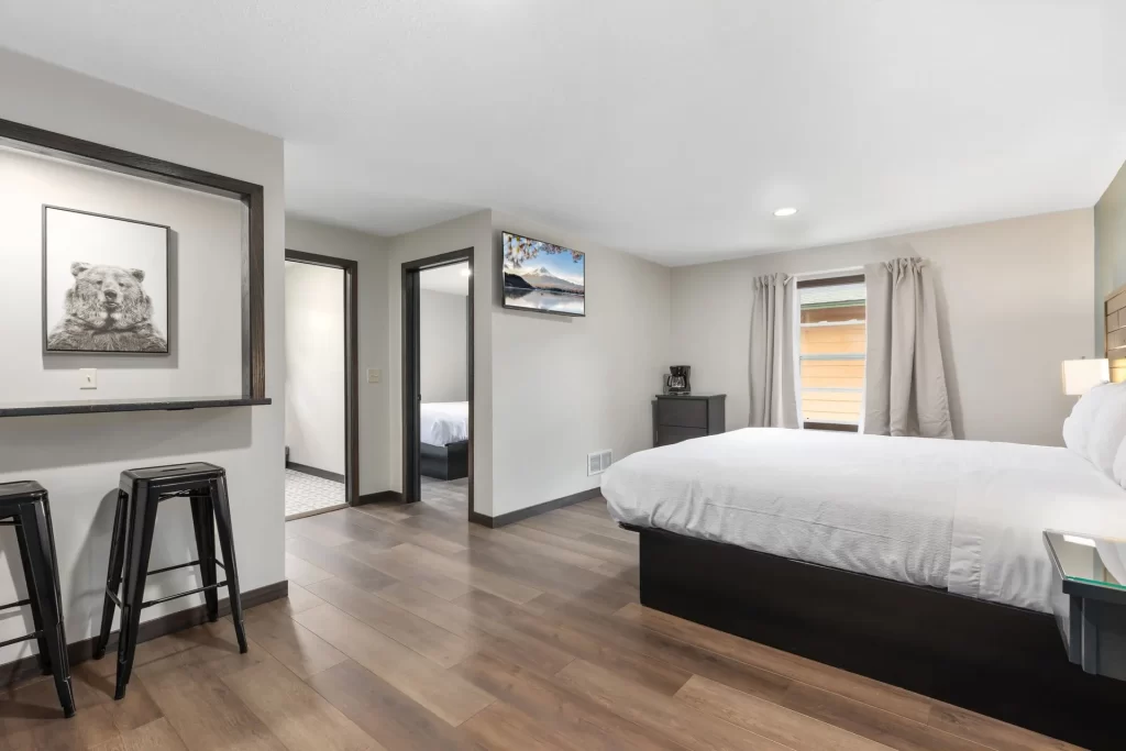 Modern bedroom with wood flooring, a large bed with white bedding, two black stools by a counter, wall art of a bear, a TV, a window with beige curtains, and a view into an adjoining room.