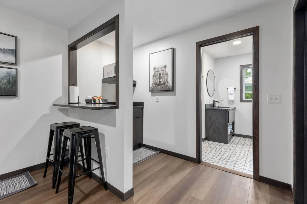 Modern apartment interior with a small kitchen area featuring a breakfast bar and three black stools, a hallway leading to a bathroom with patterned tile floor, and minimalist decor on white walls.