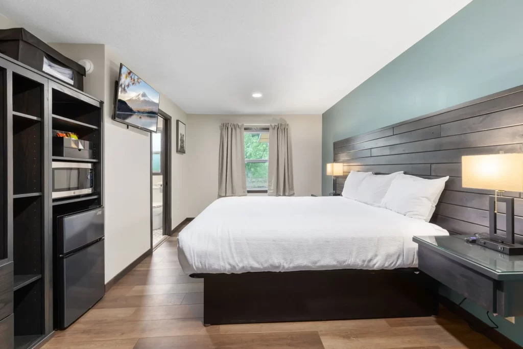 Modern hotel room with a large bed, white bedding, wooden floor, and feature wall. A flat-screen TV, shelves, microwave, and mini fridge are on the left. Window with light curtains lets in natural light.