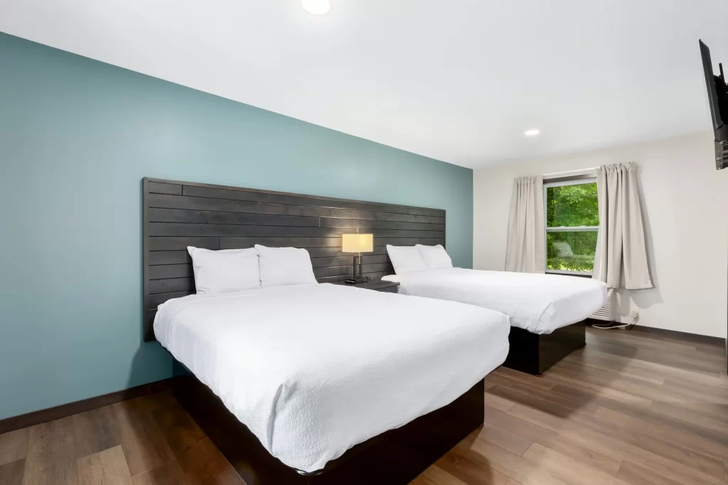 A hotel room with two queen beds, white bedding, a wooden headboard, a nightstand with a lamp, wooden flooring, a window with beige curtains, and a wall-mounted TV.
