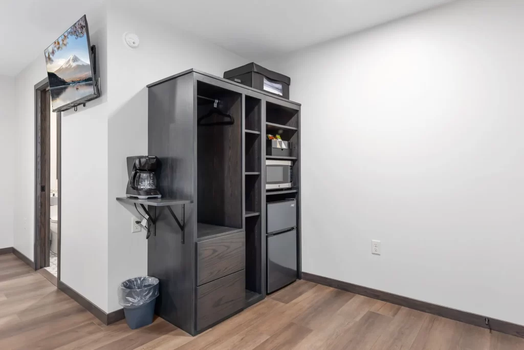 A compact kitchenette area with a dark wood wardrobe, mini fridge, microwave, coffee maker on a small shelf, trash bin, and a flat-screen TV mounted on a white wall above wood flooring.