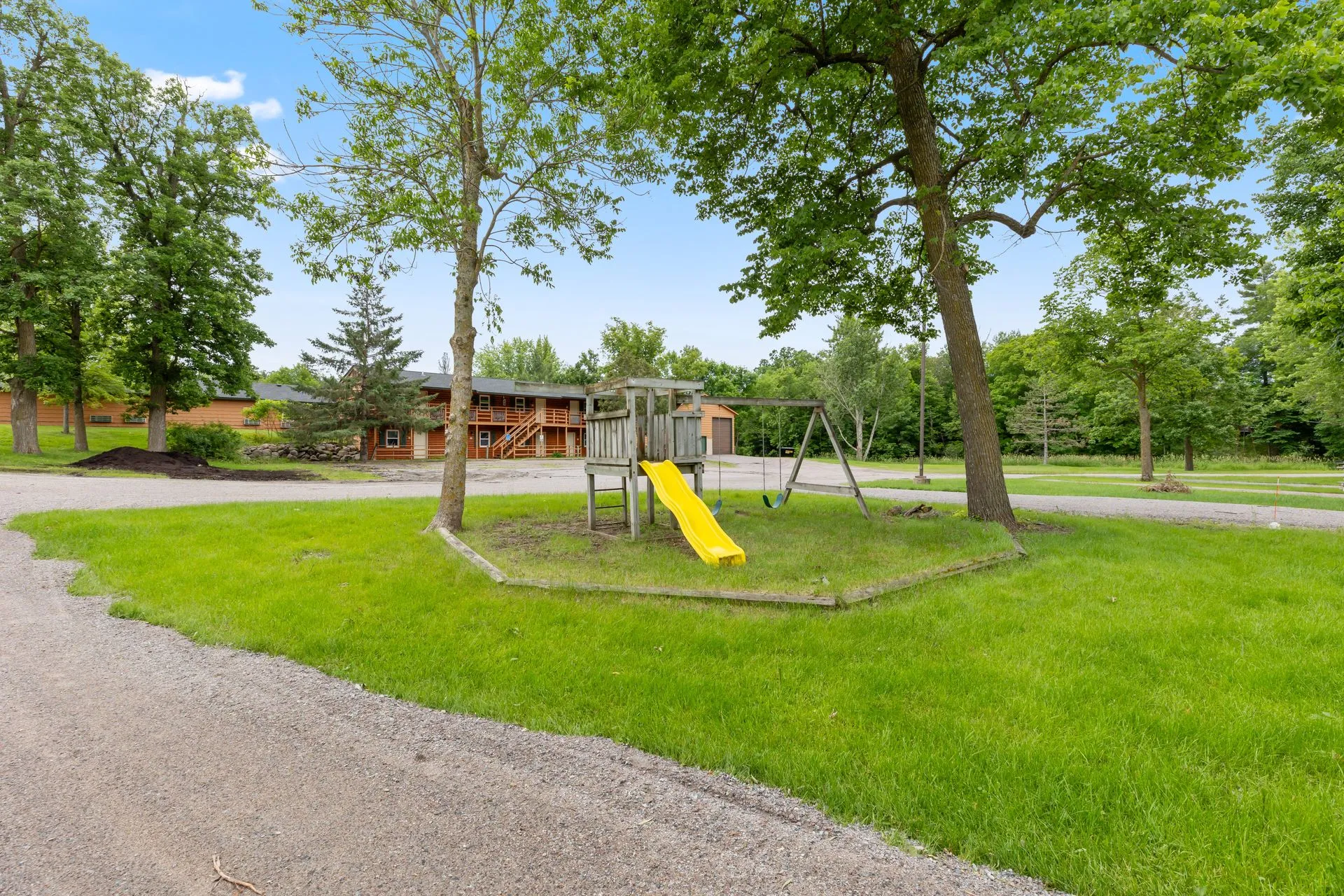 A small playground with a yellow slide and swings is set among green grass and trees, with a large house and other buildings visible in the background under a blue sky.
