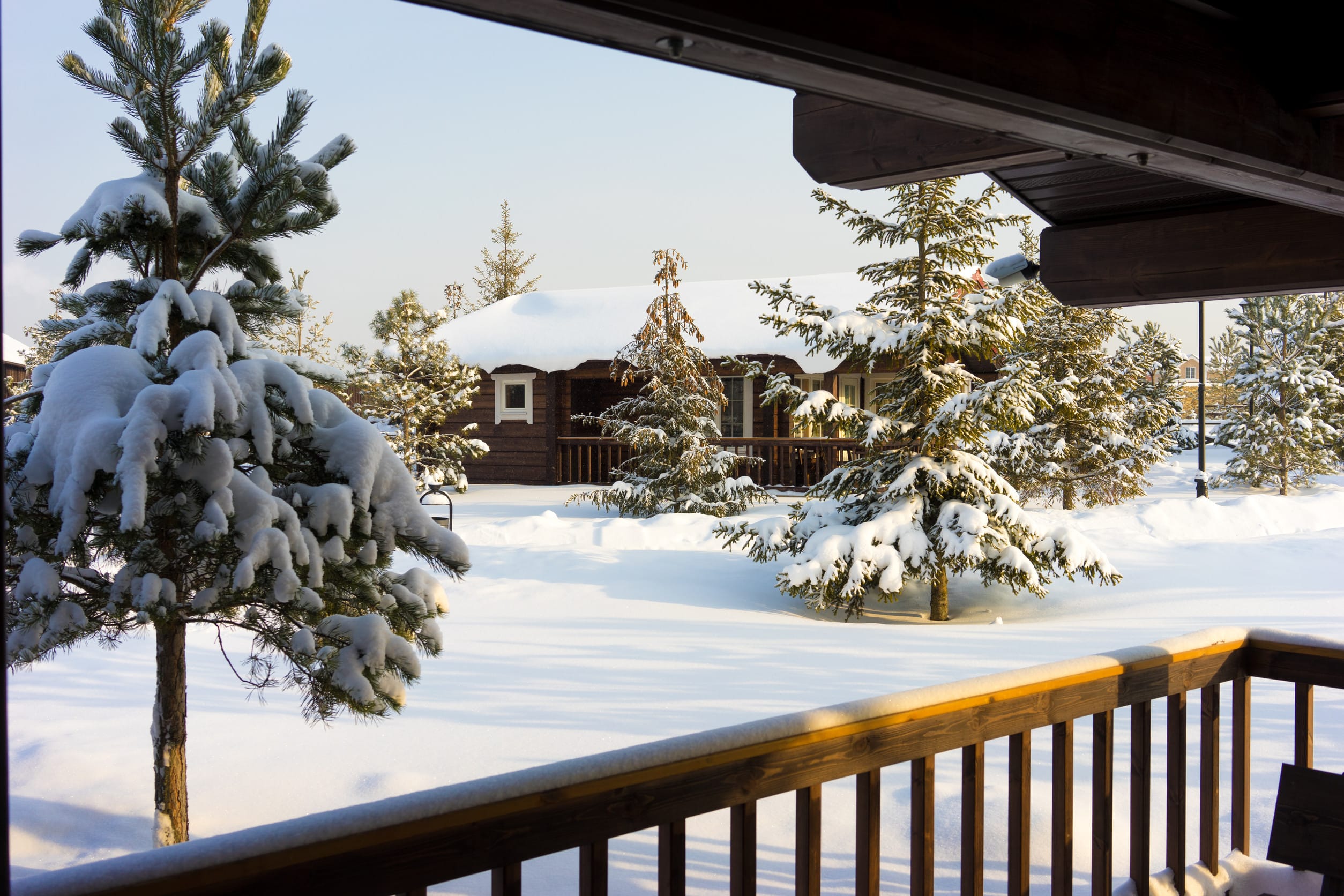 A wooden cabin surrounded by snow-covered pine trees, viewed from a porch with a wooden railing. The ground and trees are blanketed in fresh white snow under a clear sky.