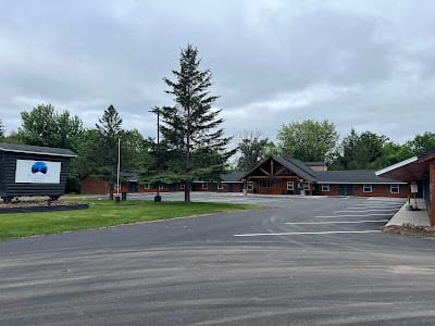 A wide, single-story building with a log cabin style and a sloped roof sits behind an empty, paved parking lot. Trees surround the area, and a sign stands to the left of the entrance. The sky is overcast.