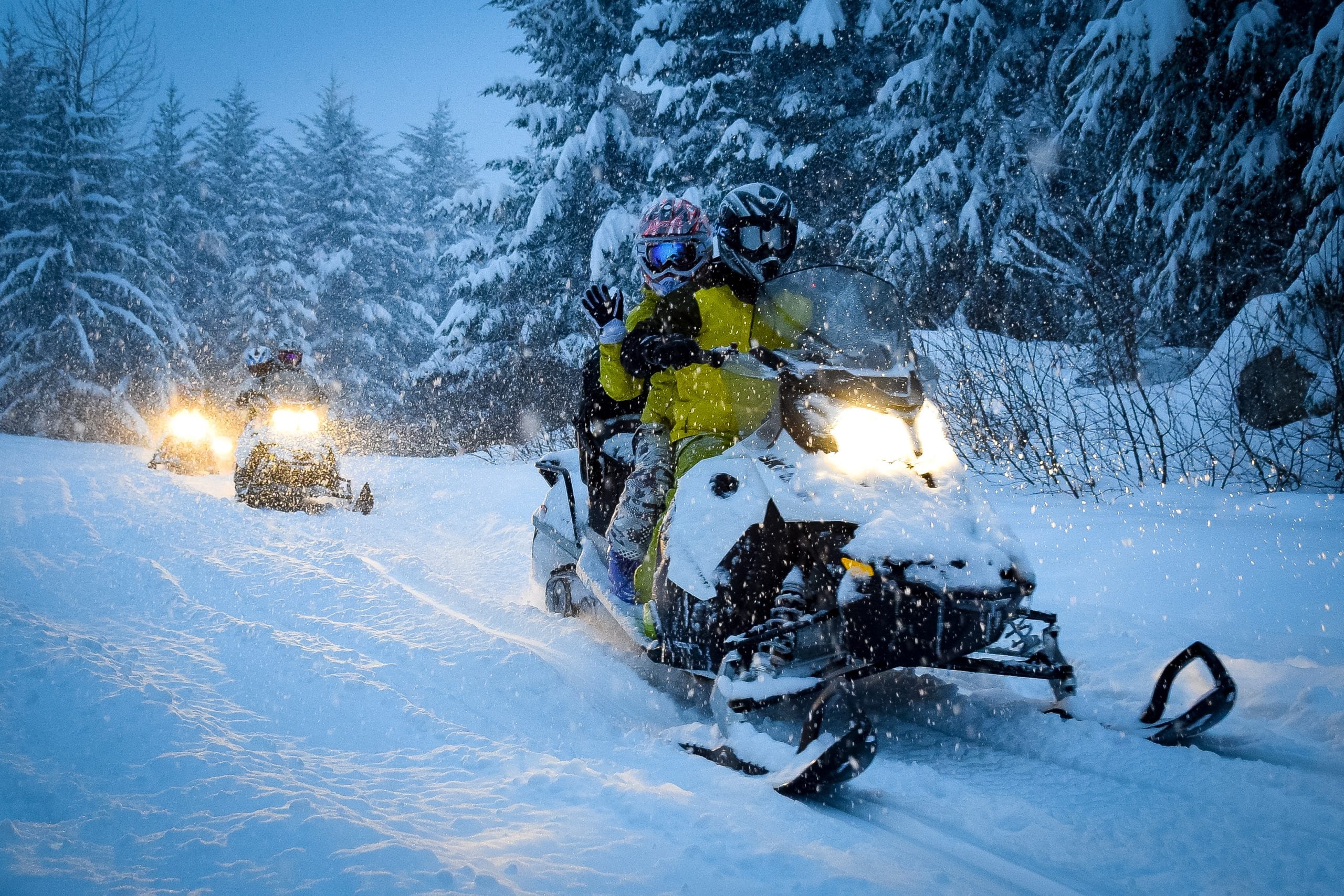 Two people ride a snowmobile through a snowy Aitkin forest, with two more snowmobiles following behind. Snow is falling heavily as all riders, dressed in winter gear, enjoy a thrilling winter getaway beneath the bright headlights.
