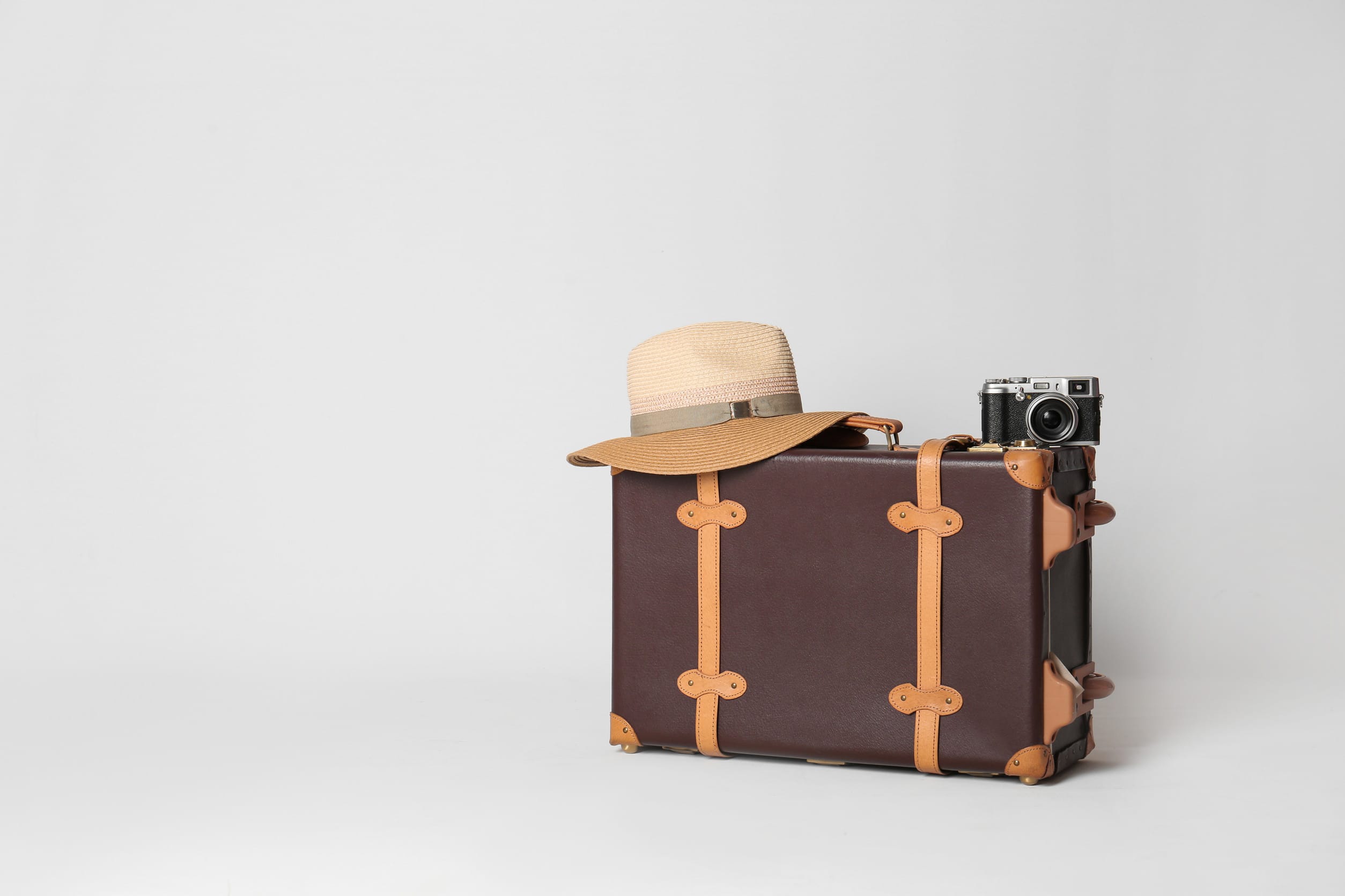 A brown vintage suitcase with tan straps sits on a light surface. A beige straw hat and an old-fashioned camera rest on top, all set against a plain white background.