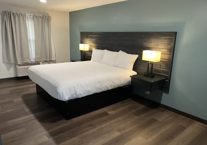 A modern bedroom features a large bed with white bedding, three pillows, a dark wooden headboard, two bedside tables with lamps, gray walls, wood flooring, a window with gray curtains, and an air conditioning unit.