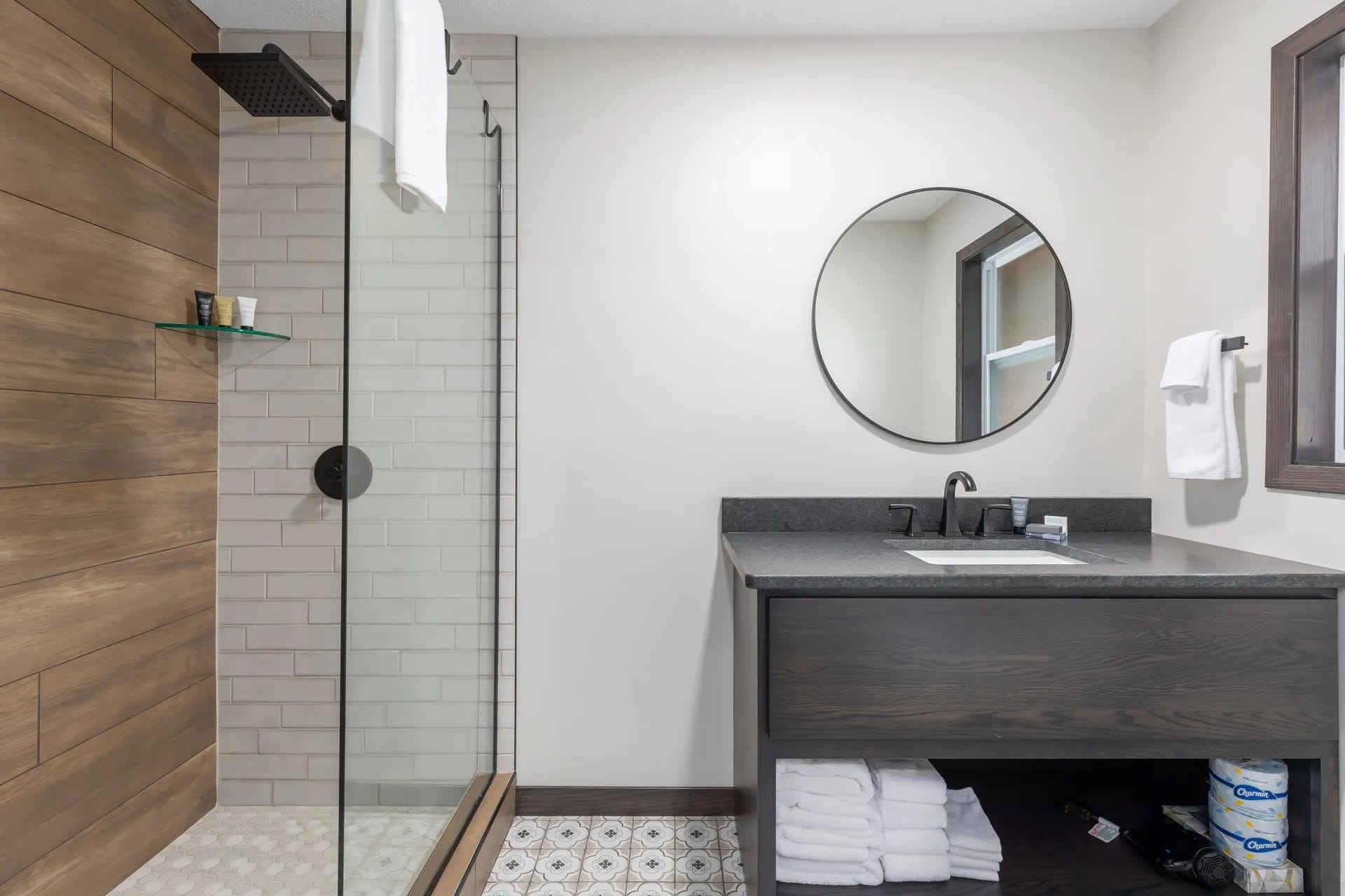 Modern bathroom with hotel amenities, a glass-enclosed shower, wood-style and white tile walls, a round mirror above a dark vanity with black faucet, folded towels on shelves, and a patterned tile floor—perfect for your Northwoods getaway.
