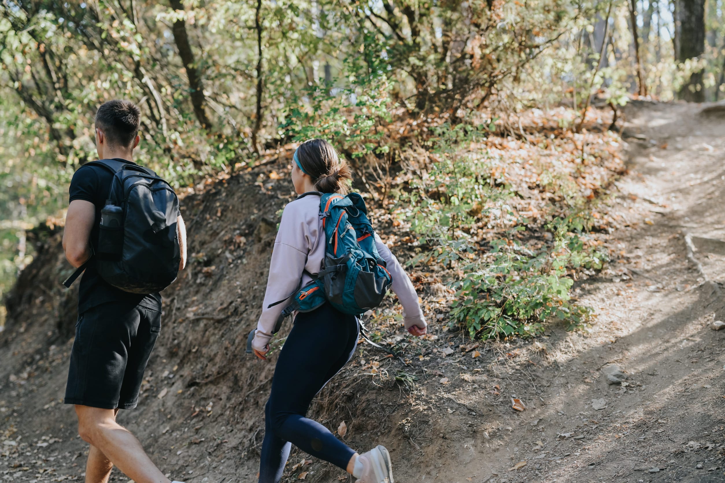 Two people with backpacks hike up a sunlit forest trail near Riverview Lodge, surrounded by trees and greenery. The person on the left wears black clothes, while the other sports a light jacket and leggings, enjoying outdoor activities together.