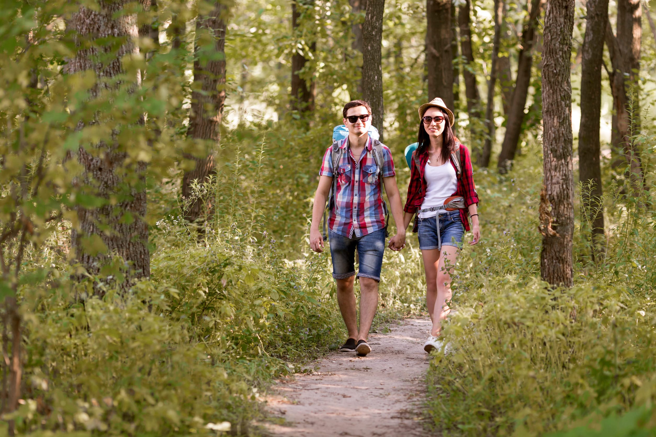 A smiling couple wearing sunglasses, shorts, and plaid shirts walks hand in hand along a forest trail surrounded by green trees and foliage on a sunny Spring weekend escape in Aitkin MN.