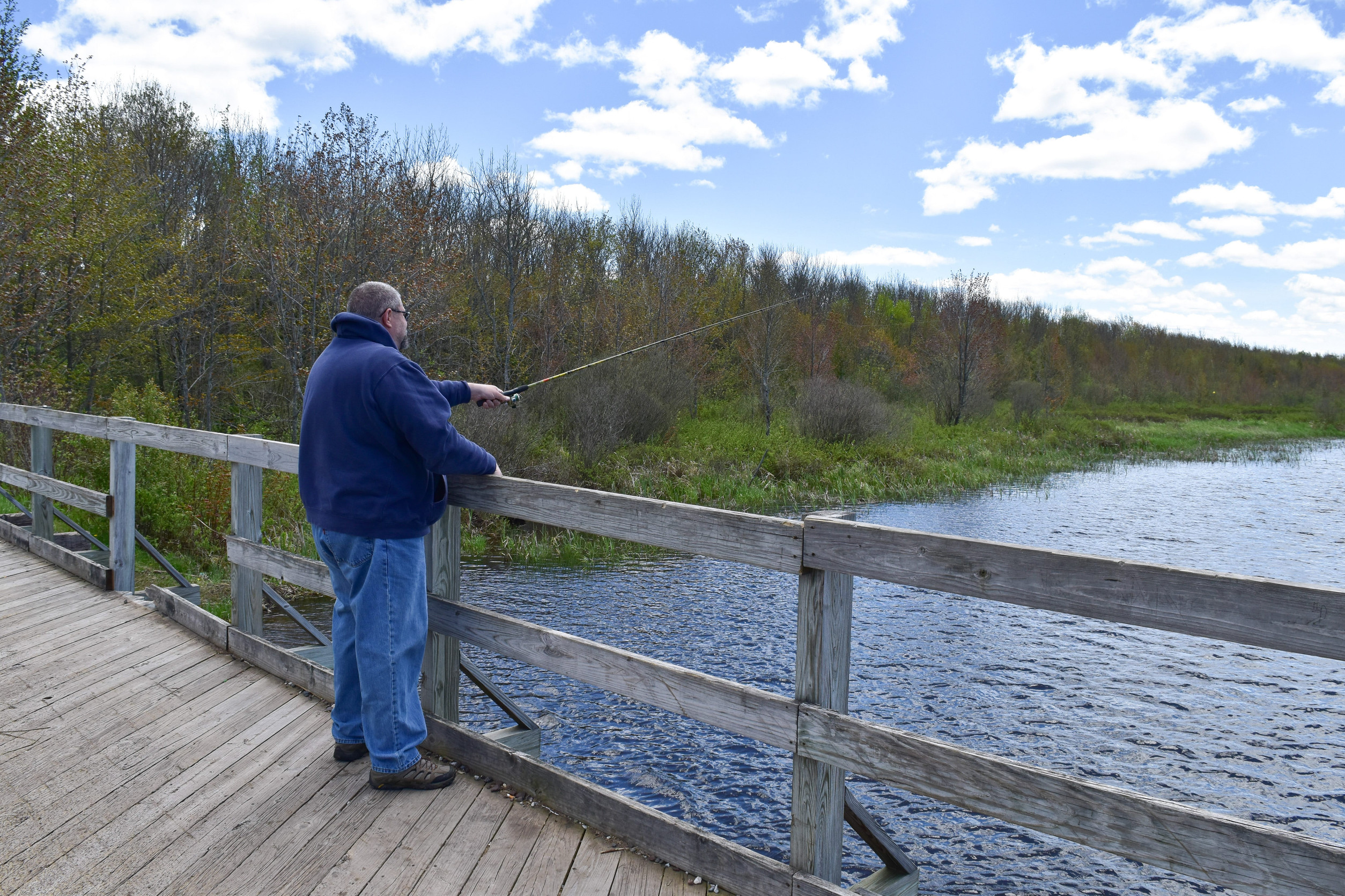 Man in a blue hoodie fishing from a wooden boardwalk beside a lake with trees and a blue sky above.
