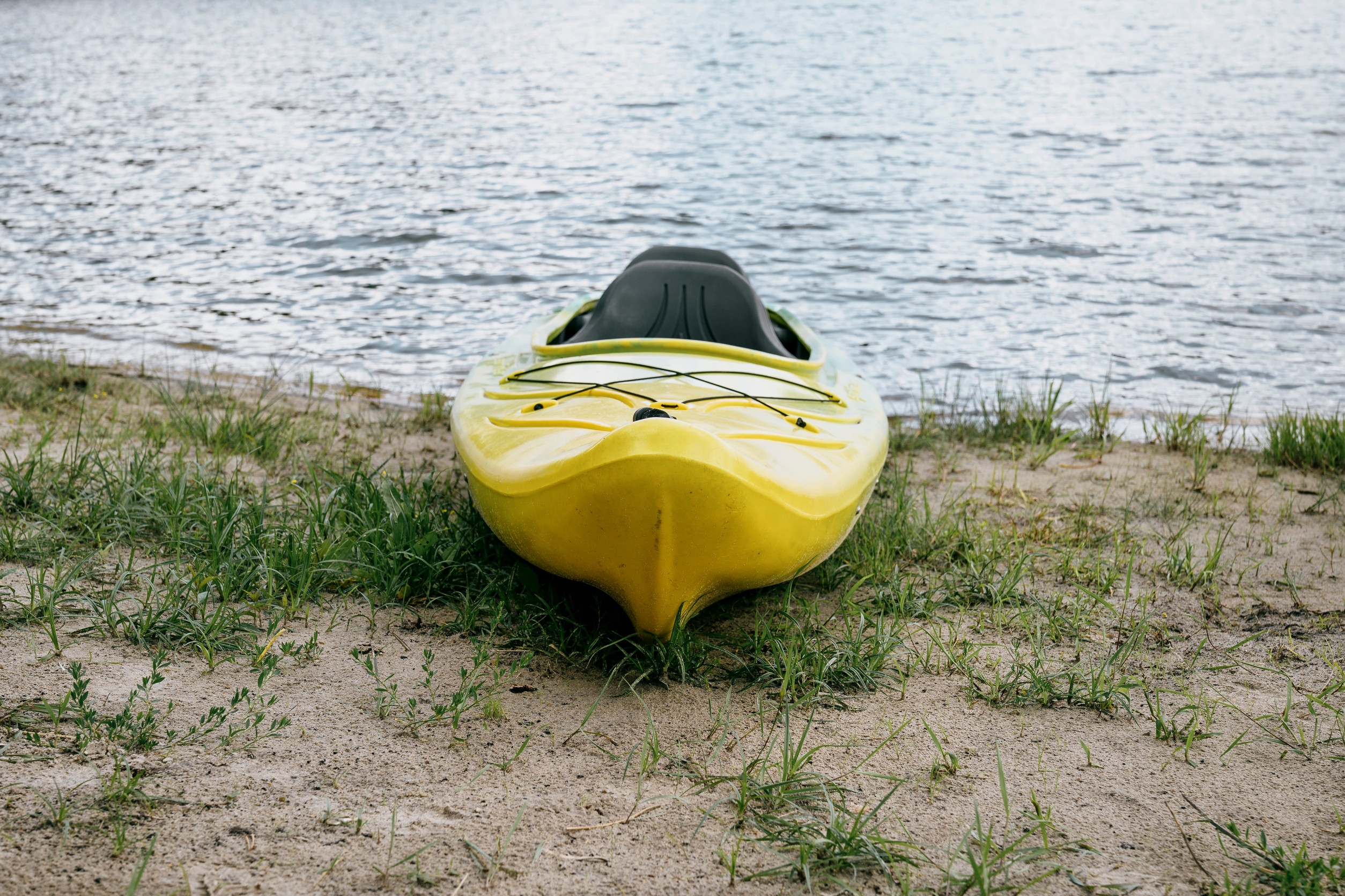 A yellow kayak rests on sandy ground by the edge of a calm lake in Aitkin, with some green grass around it and water in the background—an inviting spot for kayaking enthusiasts to begin their adventure.