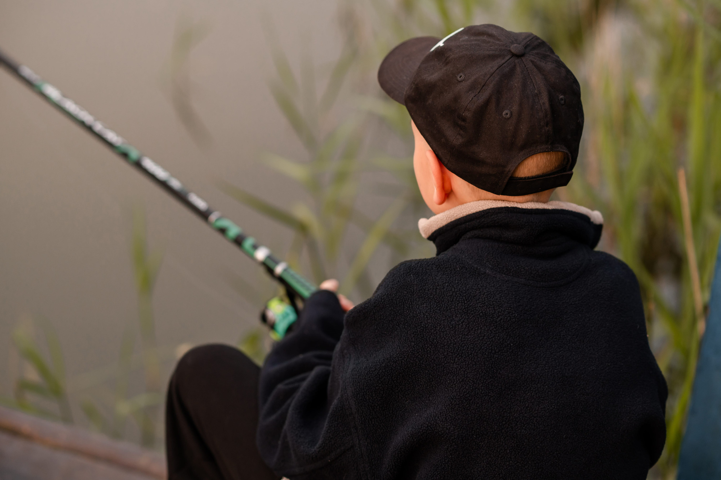 A child wearing a black cap and jacket sits by the water holding a fishing rod, facing away from the camera. Green reeds and calm water are visible in the background, capturing the peaceful atmosphere of family fishing lakes near Aitkin.