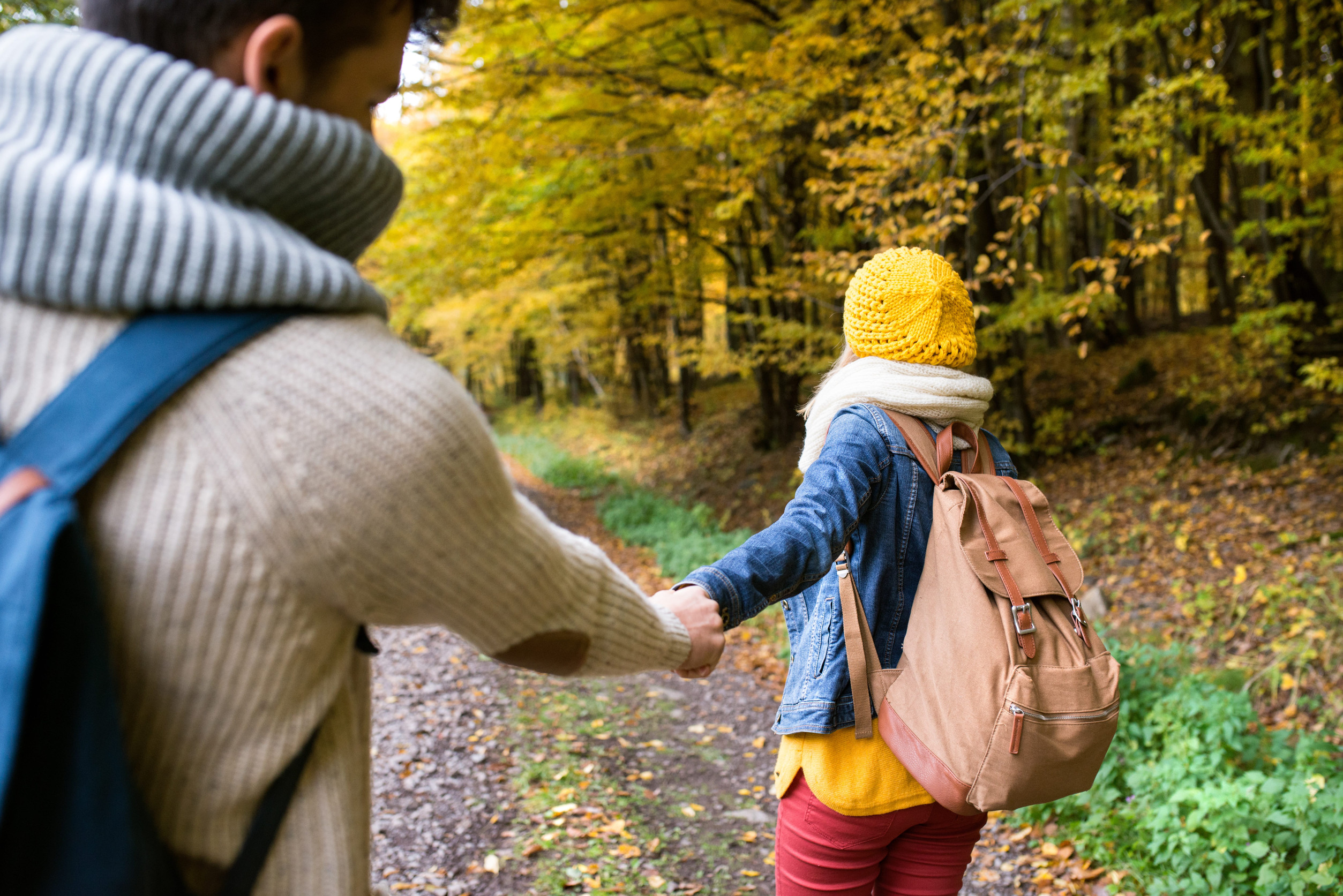 A man in a knit sweater and scarf leads a woman wearing a yellow beanie and backpack by the hand along a forest path on an autumn vacation.